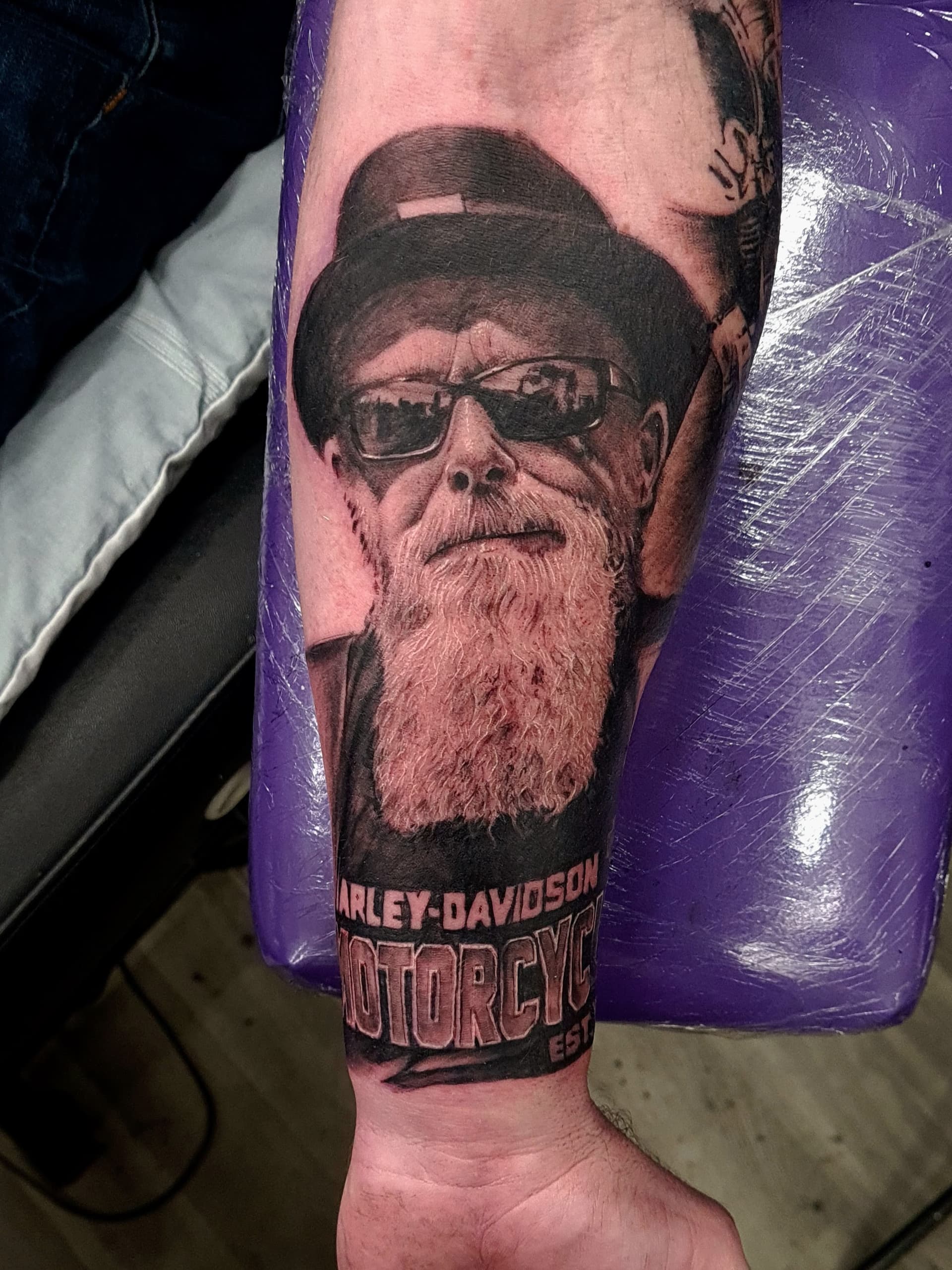 Detailed black and grey realism tattoo of a man with glasses and a beard on forearm.