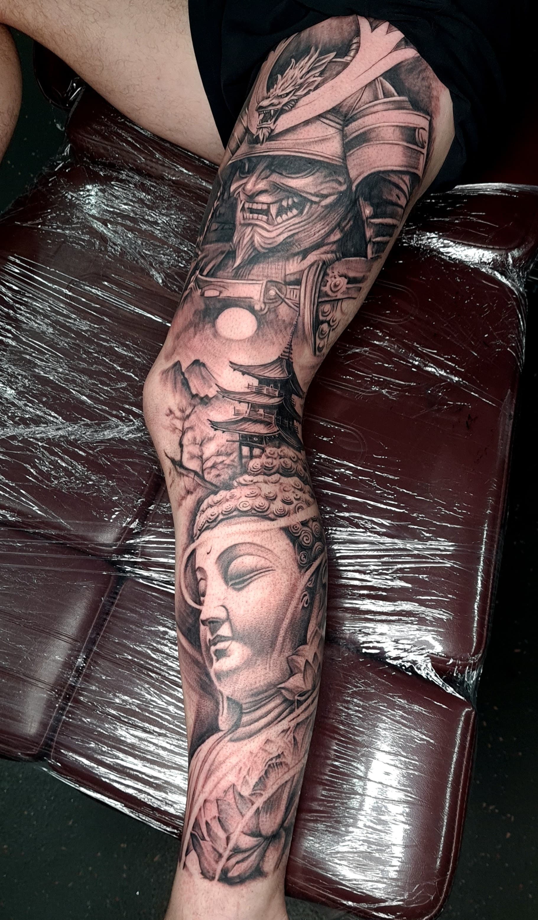 Detailed black grey realism tattoo of a samurai and buddha on the leg.