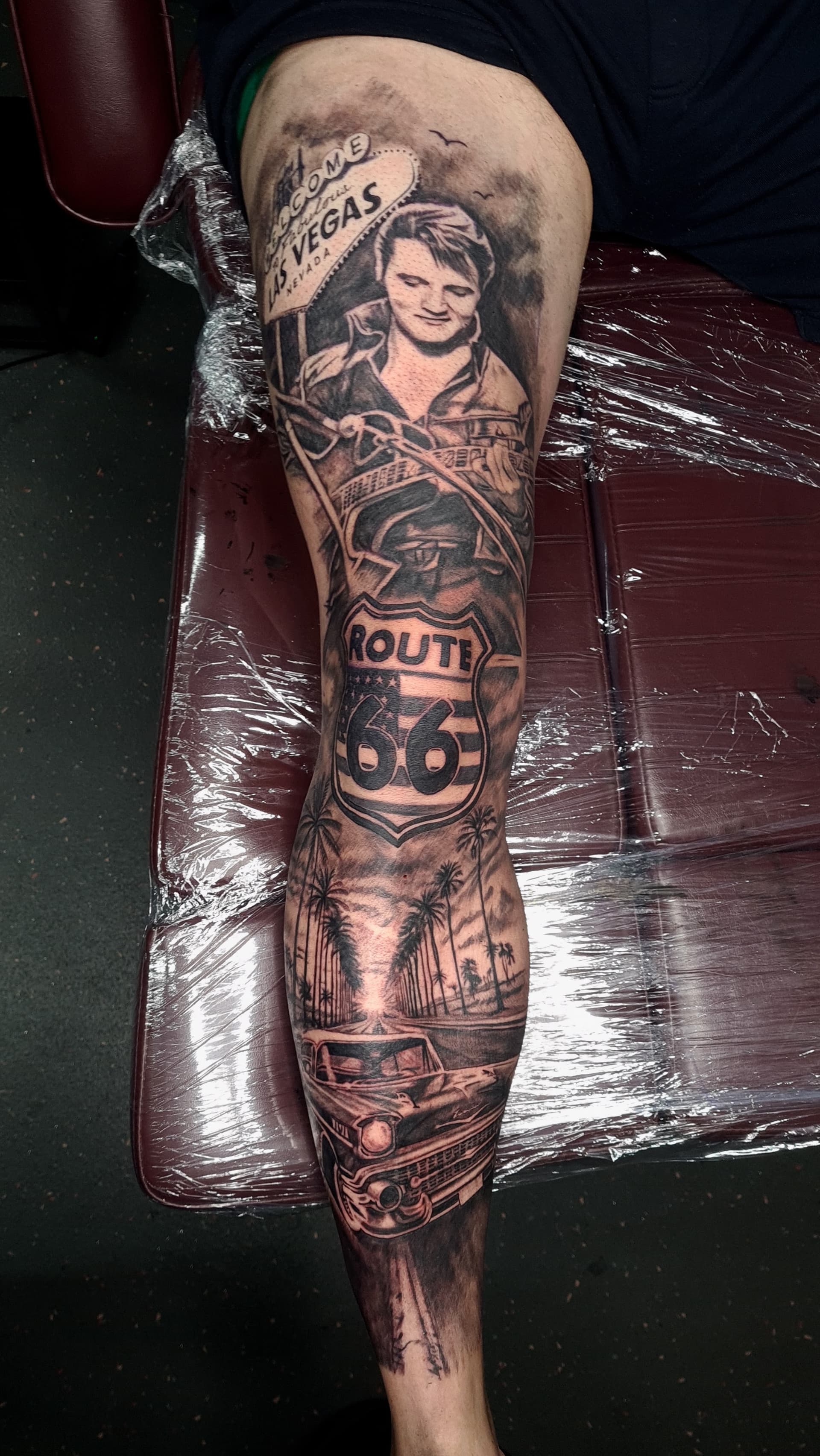 Detailed black and grey realism tattoo featuring Elvis and Route 66 scenery on leg.