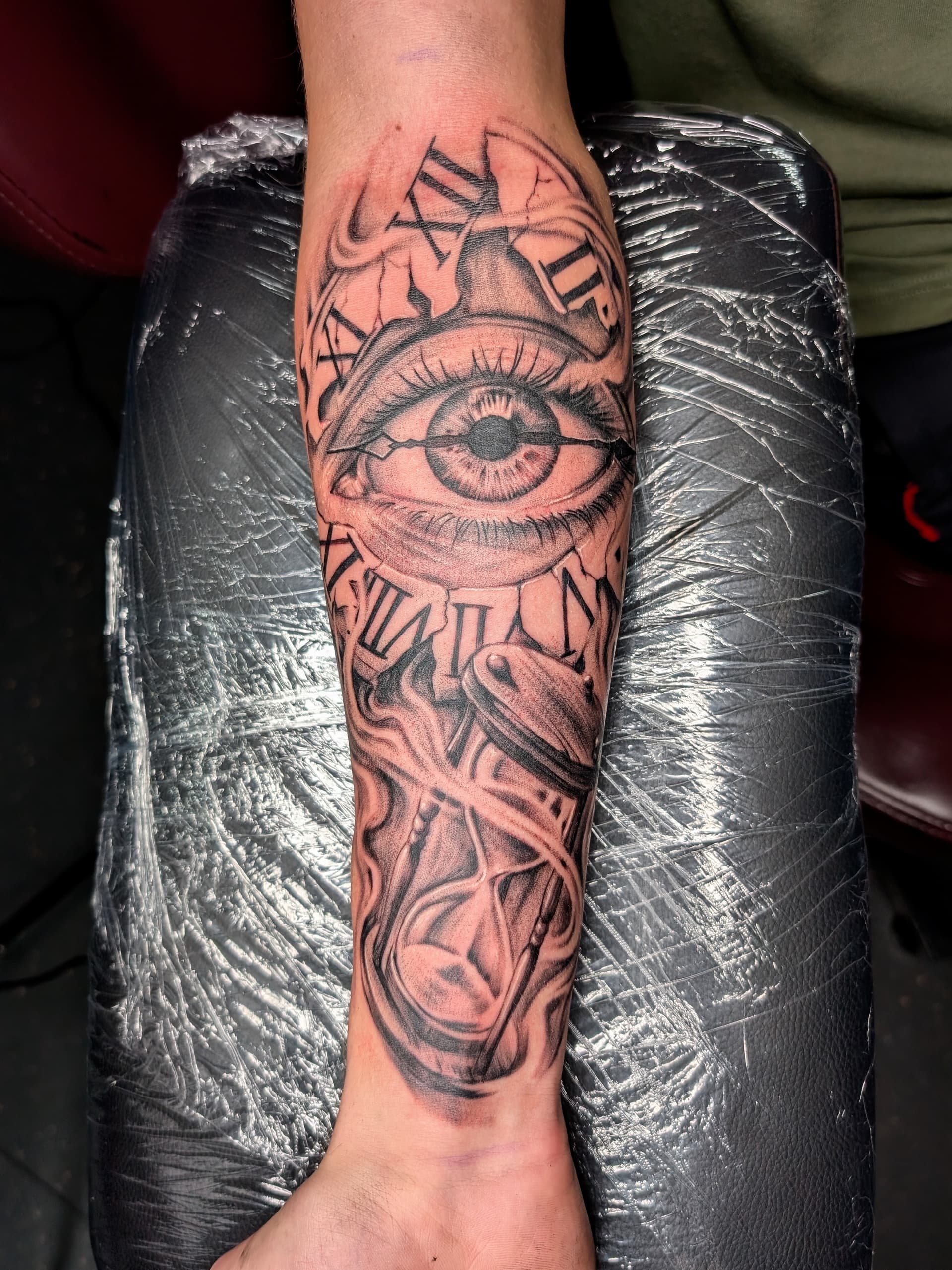 Intricate black and grey realism tattoo of an eye and hourglass on forearm.