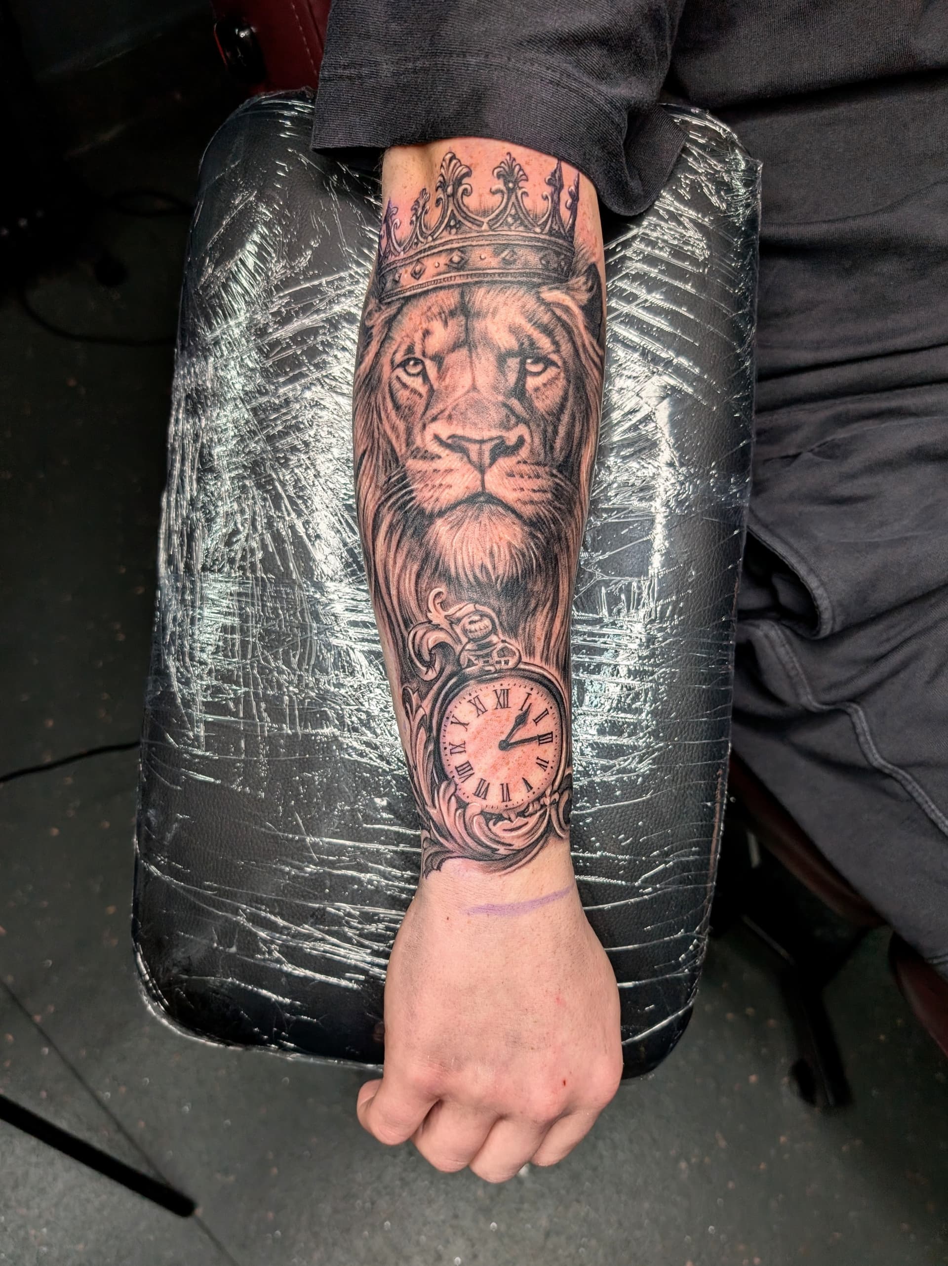 Detailed black and grey realism tattoo featuring a lion and clock on forearm.