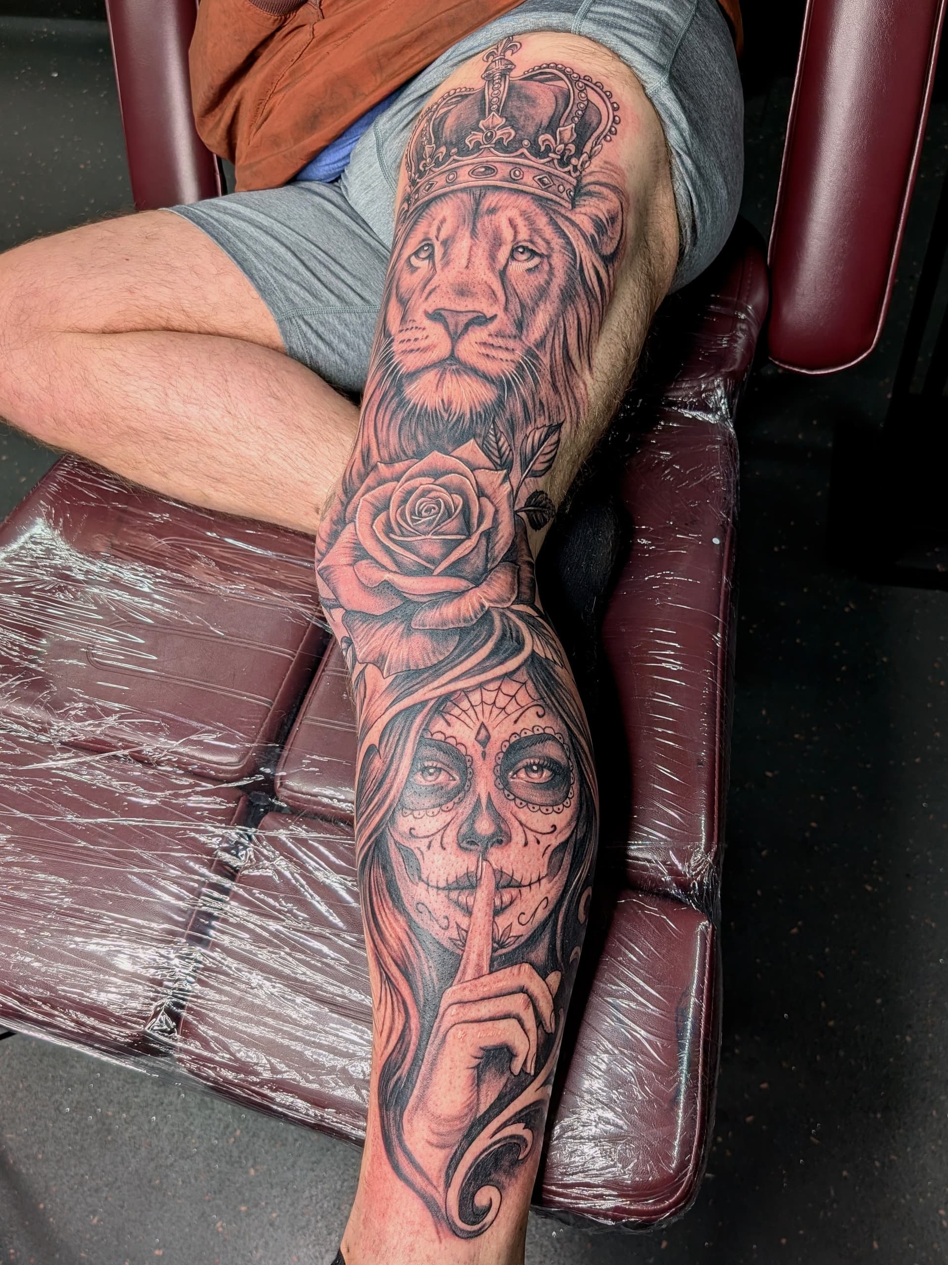 Tattoo of a lion with a crown and a skull woman with a rose on leg.