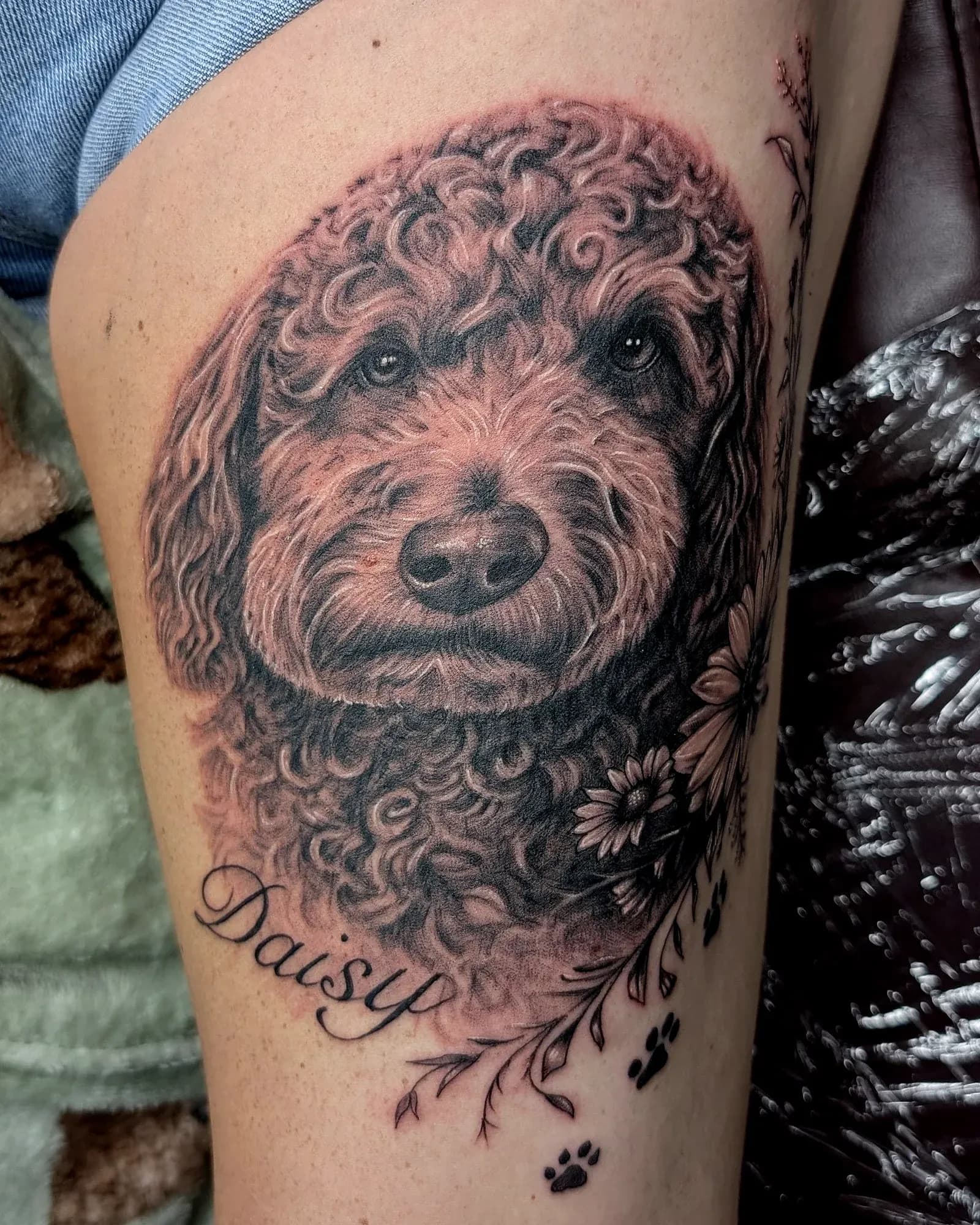 Black and grey realism tattoo of a golden doodle with daisies, her name and paw prints on the thigh.