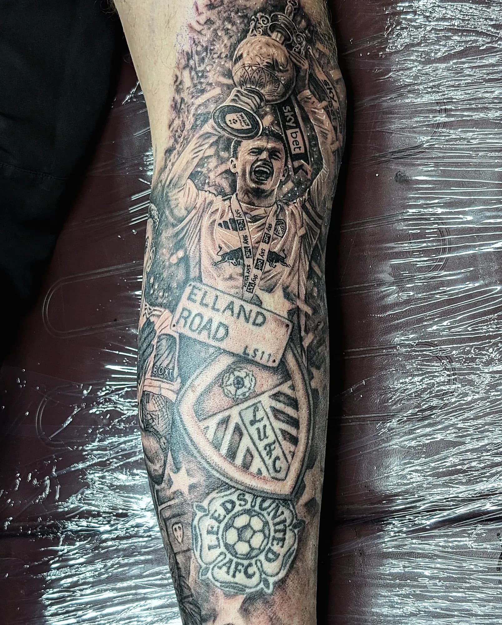Black and grey trophy-lift tattoo above an Elland Road sign, worked around an older Leeds badge done by another artist.