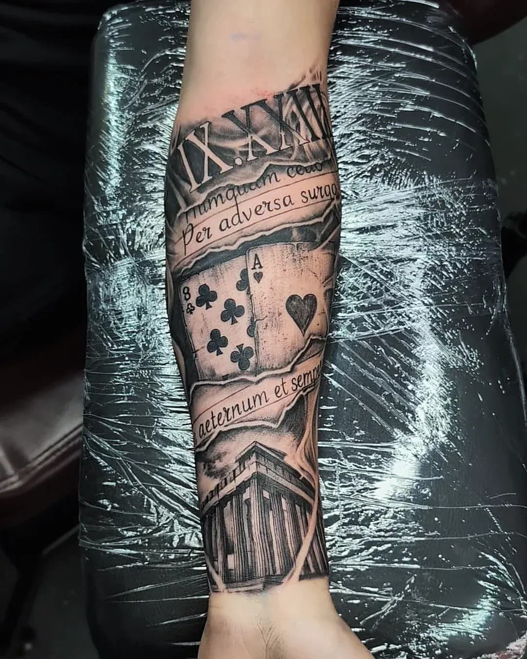 Black and grey realism tattoo of two playing cards, torn-paper Latin script and a Greek temple on the inner forearm.