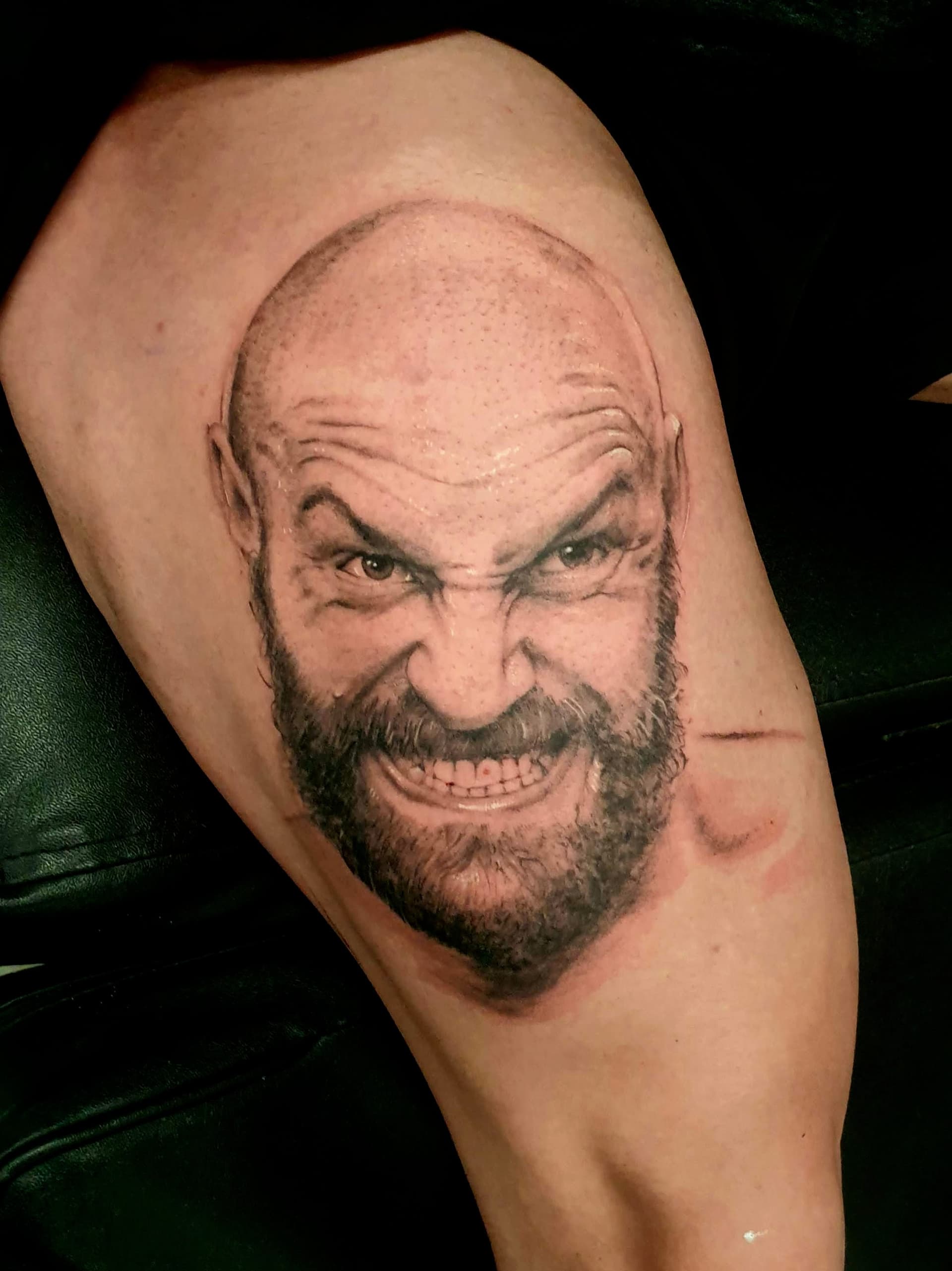 Black and grey realism tattoo of a snarling bald bearded man's face on the upper arm